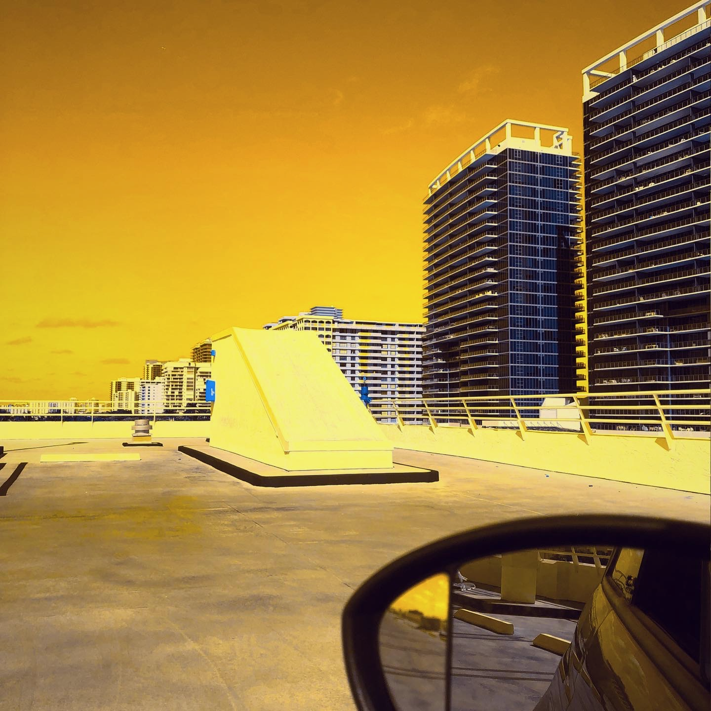 Open rooftop parking garage overlooking skyscrapers. Camera captured the driver-side’s mirror—yellow aesthetic.